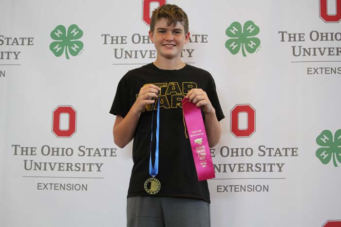 Boy wearing a Star Wars shirt stands at an OSU Extension backdrop, holding a gold medal with a blue ribbon and a pink award ribbon.