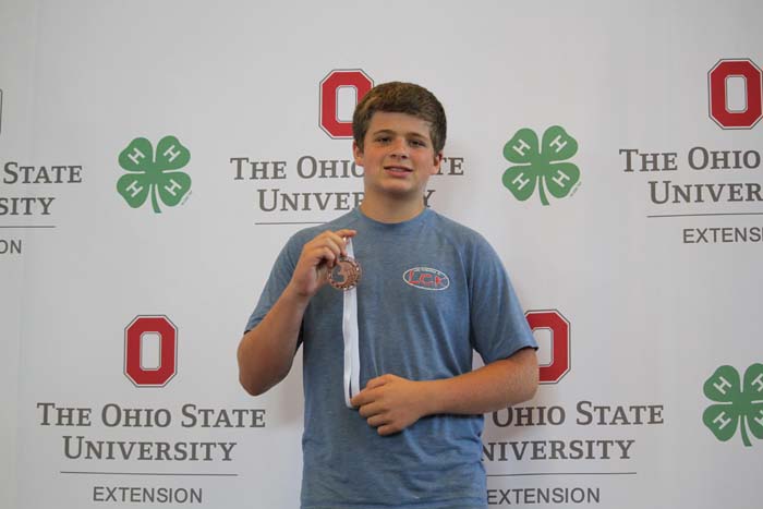 Young man in a blue shirt holding a medal with a white ribbon in front of an Ohio State University Extension backdrop.
