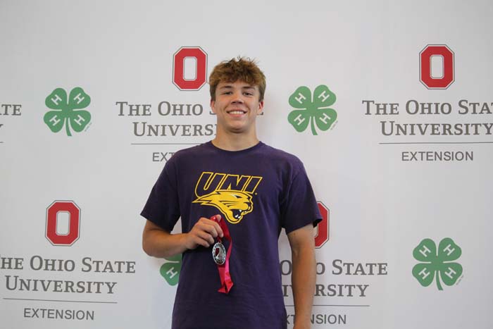 Smiling young man in a purple UNI t-shirt holds a medal with red ribbon, standing in front of The Ohio State University Extension backdrop.