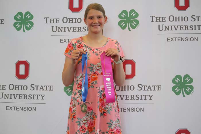 Young woman in a pink floral dress holds two award ribbons (blue and pink) in front of a backdrop that reads The Ohio State University Extension and features OSU logos.