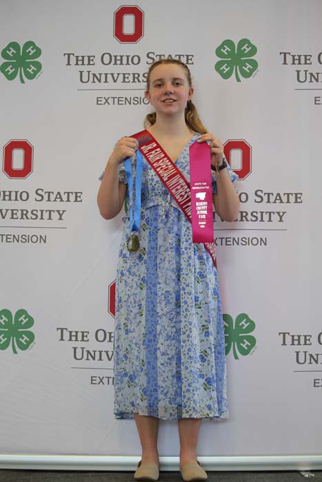Young girl in a blue floral dress holds two award ribbons and a medal in front of an Ohio State University backdrop.