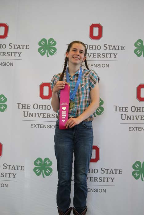 Teenage girl with braided hair in a plaid shirt and jeans posing with a pink trophy and blue lanyard, in front of an Ohio State University Extension backdrop.