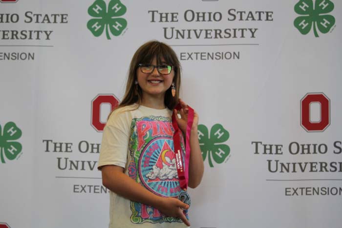 Smiling girl with glasses stands against a backdrop featuring The Ohio State University Extension logos, wearing a colorful graphic tee and holding a pink lanyard.