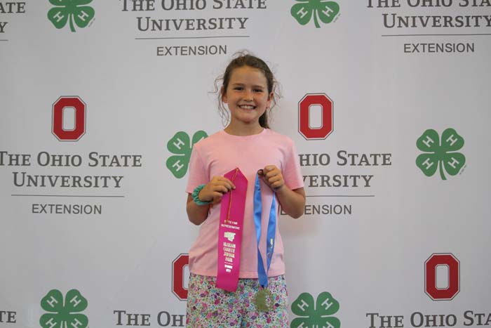 Smiling girl in a pink shirt holds pink and blue award ribbons at an Ohio State University Extension backdrop.