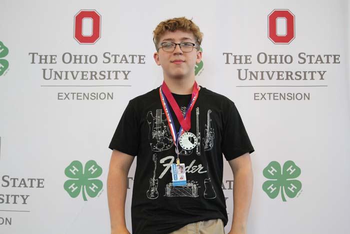 Young man with glasses wearing a black graphic t-shirt, medals around his neck, standing in front of an Ohio State University Extension backdrop featuring 4-H clover logos.