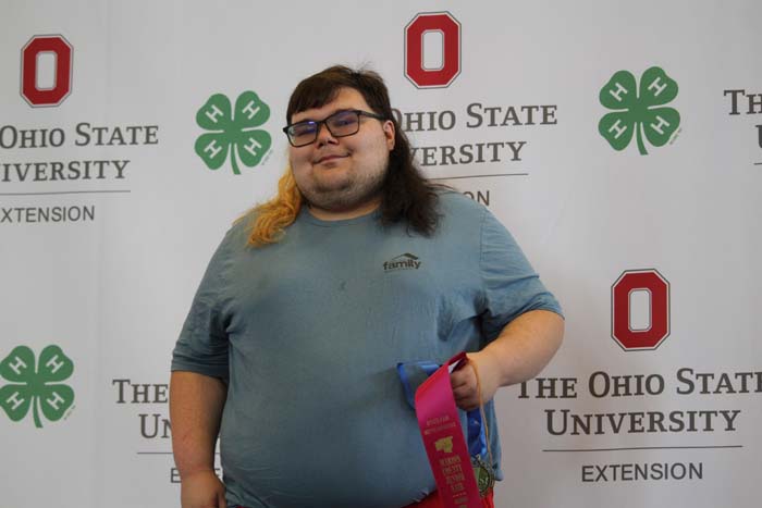 Person with glasses and long hair in a blue shirt posing in front of an Ohio State University Extension backdrop, holding a pink ribbon.