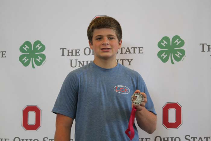 Young man in a blue T-shirt stands against a backdrop with The Ohio State University branding, holding a silver second-place medal with a pink ribbon.