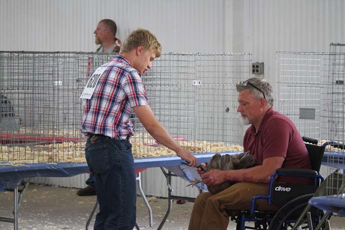 Man in a plaid shirt hands something to a man in a wheelchair holding a small dog at a table, with kennels in the background.