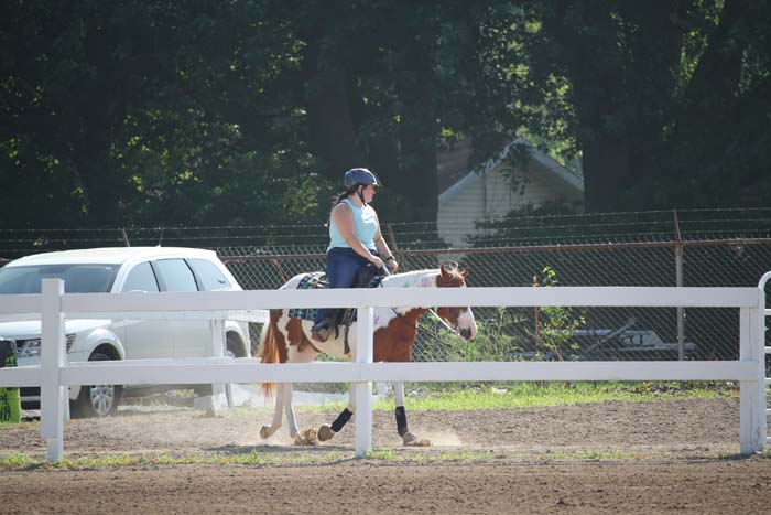 Person wearing a helmet rides a brown-and-white pony inside a white fenced riding arena outdoors.