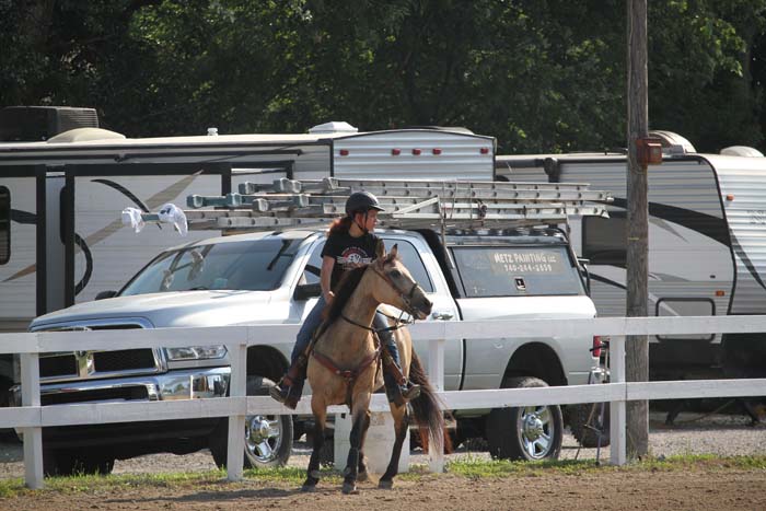 Person wearing a helmet riding a brown horse inside a fenced arena, with trucks and horse trailers parked in the background.