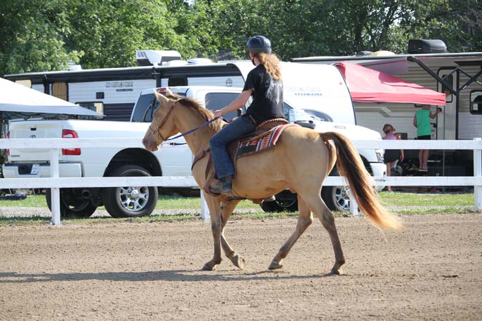 Rider wearing a helmet on a palomino horse walking in an outdoor riding arena, with horse trailers in the background.