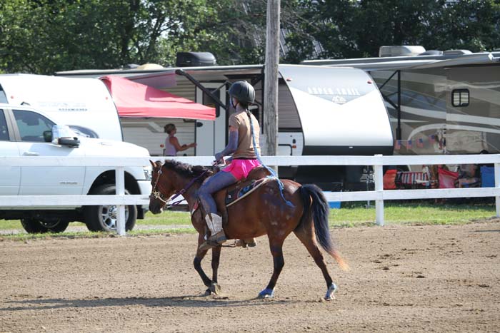 Rider wearing a helmet on a chestnut horse trotting in a sandy riding arena, with horse trailers and vehicles in the background.