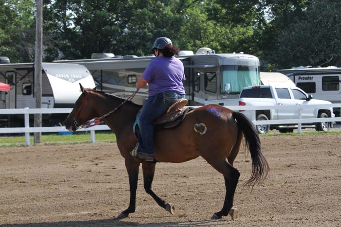 Rider wearing a purple shirt and helmet guides a brown horse across a dirt arena with trailers in the background.