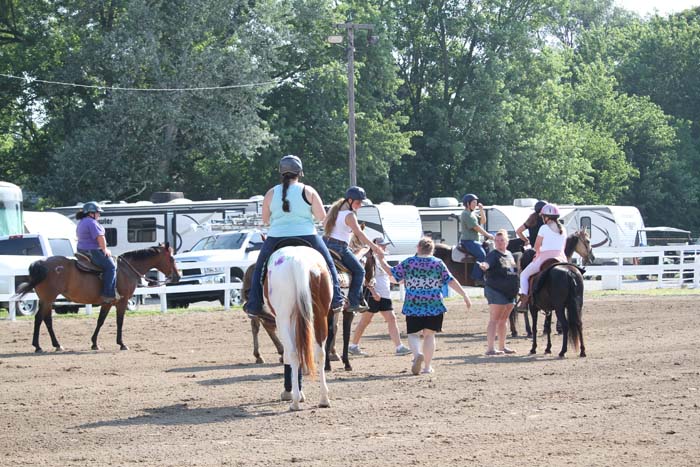 Group of people in a sunny riding arena with horses and riders, a white fence and trailers in the background.