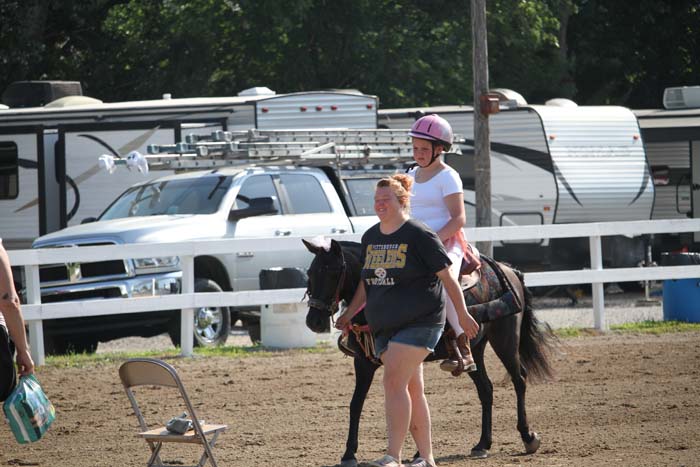 Girl in a pink helmet rides a dark pony while a woman guides it in a dirt riding arena; trailers and a white pickup truck visible in the background.