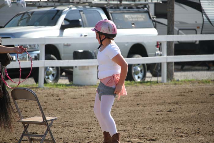 Young girl wearing a pink helmet and white shirt stands in a dirt riding arena next to a chair, with a fence and vehicles in the background.
