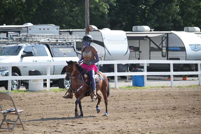 Rider wearing a helmet on a brown horse trotting in a dirt arena; trailers and vehicles visible behind the fence.