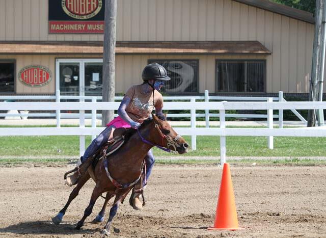 Rider in helmet guiding a brown horse around an orange cone in a dirt arena.