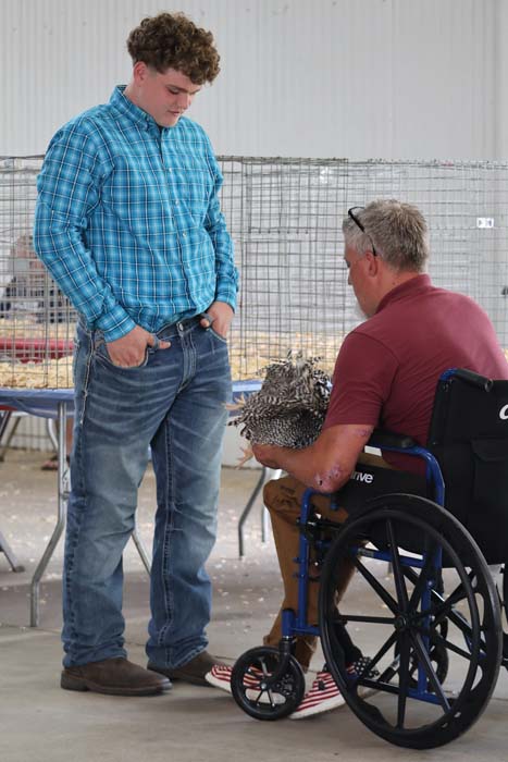 Two men talk in an indoor event space; one standing in a blue plaid shirt, the other in a wheelchair holding a spiky animal.