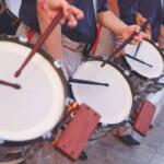 Row of street drummers in a parade, hitting white drumheads with sticks and mallets in motion