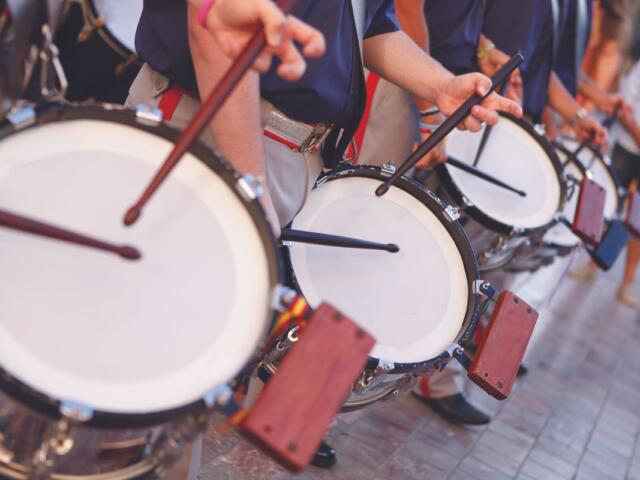Row of street drummers in a parade, hitting white drumheads with sticks and mallets in motion
