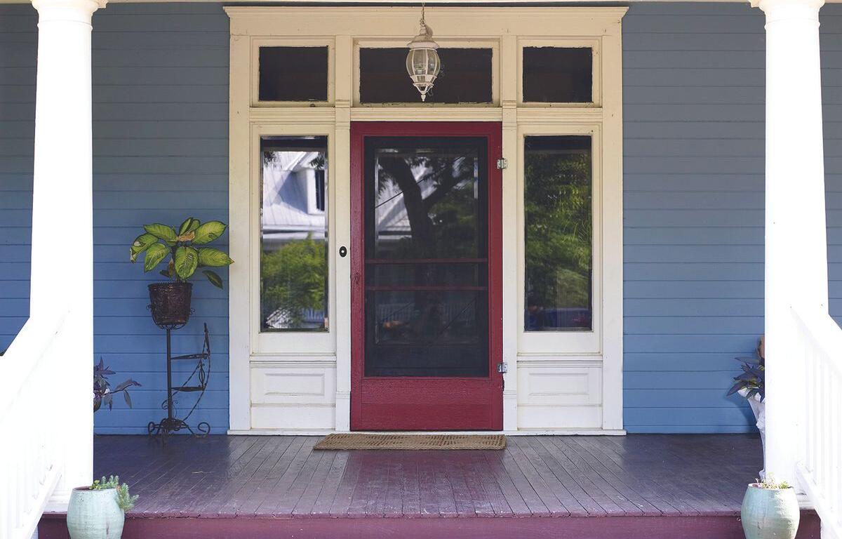 Front porch of a blue house with white trim and a red storm door, flanked by windows and potted plants on the deck area.
