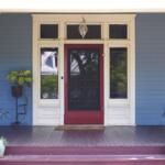 Front porch of a blue house with white trim and a red storm door, flanked by windows and potted plants on the deck area.