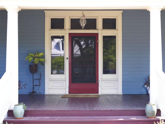 Front porch of a blue house with white trim and a red storm door, flanked by windows and potted plants on the deck area.