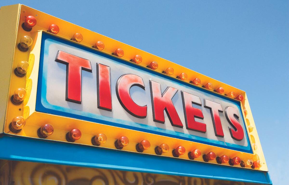 Bright yellow carnival ticket sign with illuminated bulbs and the word TICKETS against a blue sky.