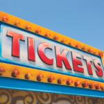 Bright yellow carnival ticket sign with illuminated bulbs and the word TICKETS against a blue sky.