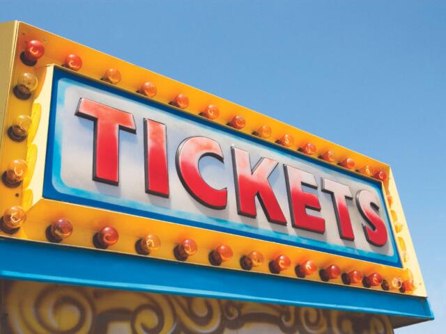 Bright yellow carnival ticket sign with illuminated bulbs and the word TICKETS against a blue sky.