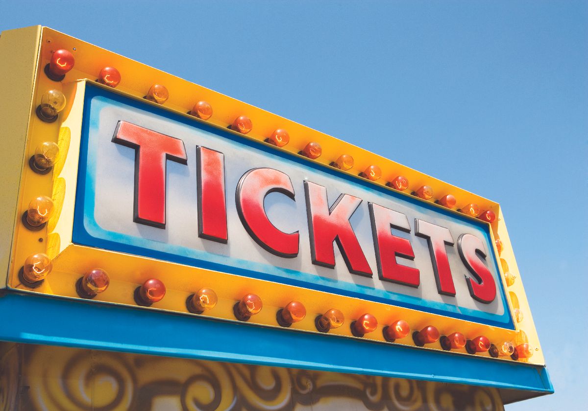 Bright yellow carnival ticket sign with illuminated bulbs and the word TICKETS against a blue sky.