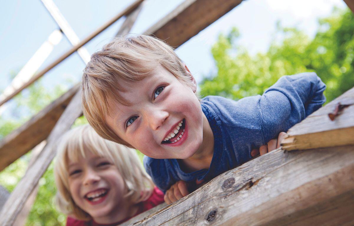 Two young children lean over a weathered wooden railing outdoors, smiling and enjoying playtime.