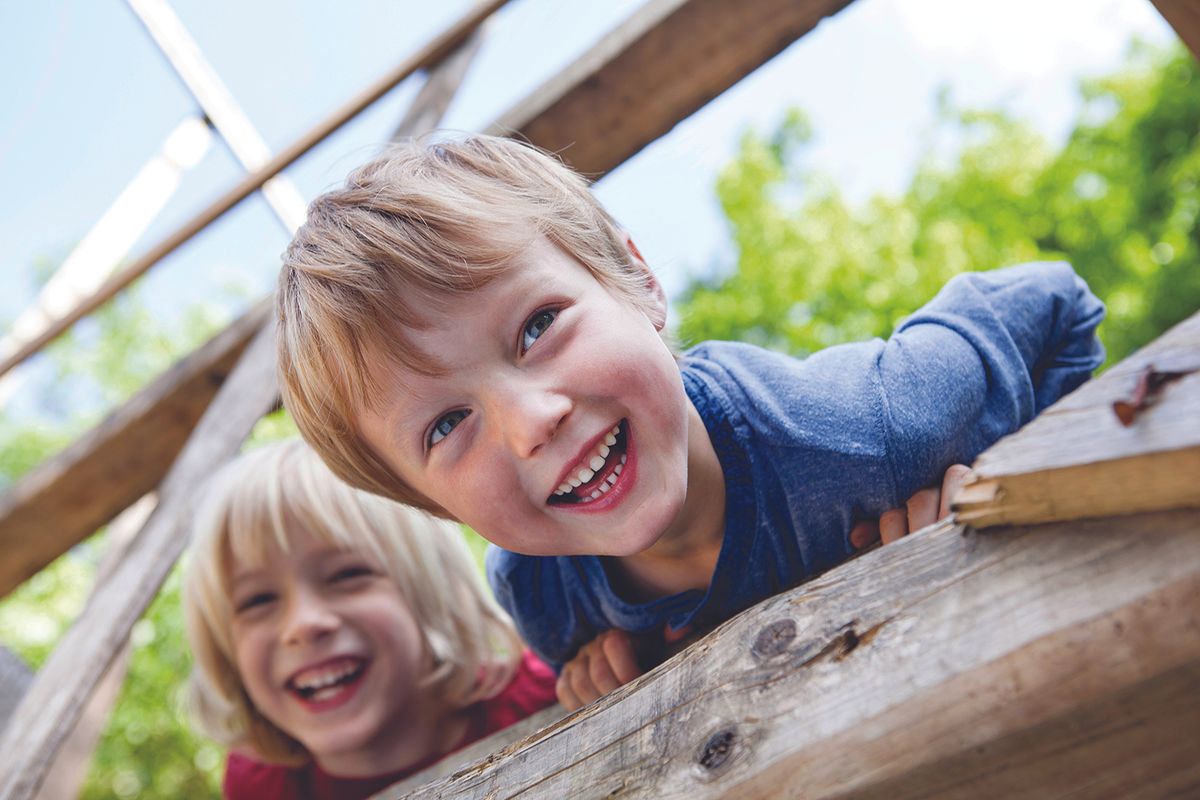 Two young children lean over a weathered wooden railing outdoors, smiling and enjoying playtime.