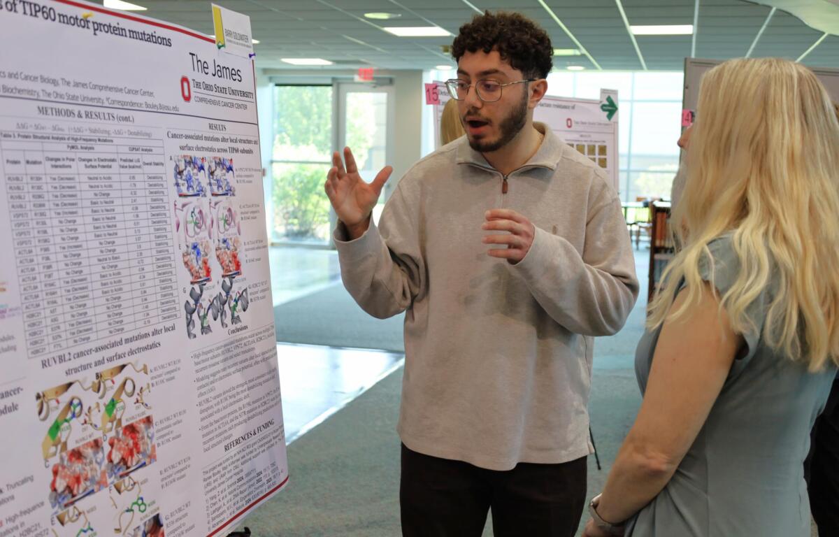 Man explains a scientific poster about cancer-related mutations to a woman at a conference or poster session.
