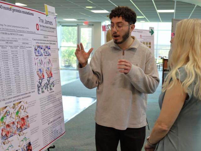 Man explains a scientific poster about cancer-related mutations to a woman at a conference or poster session.