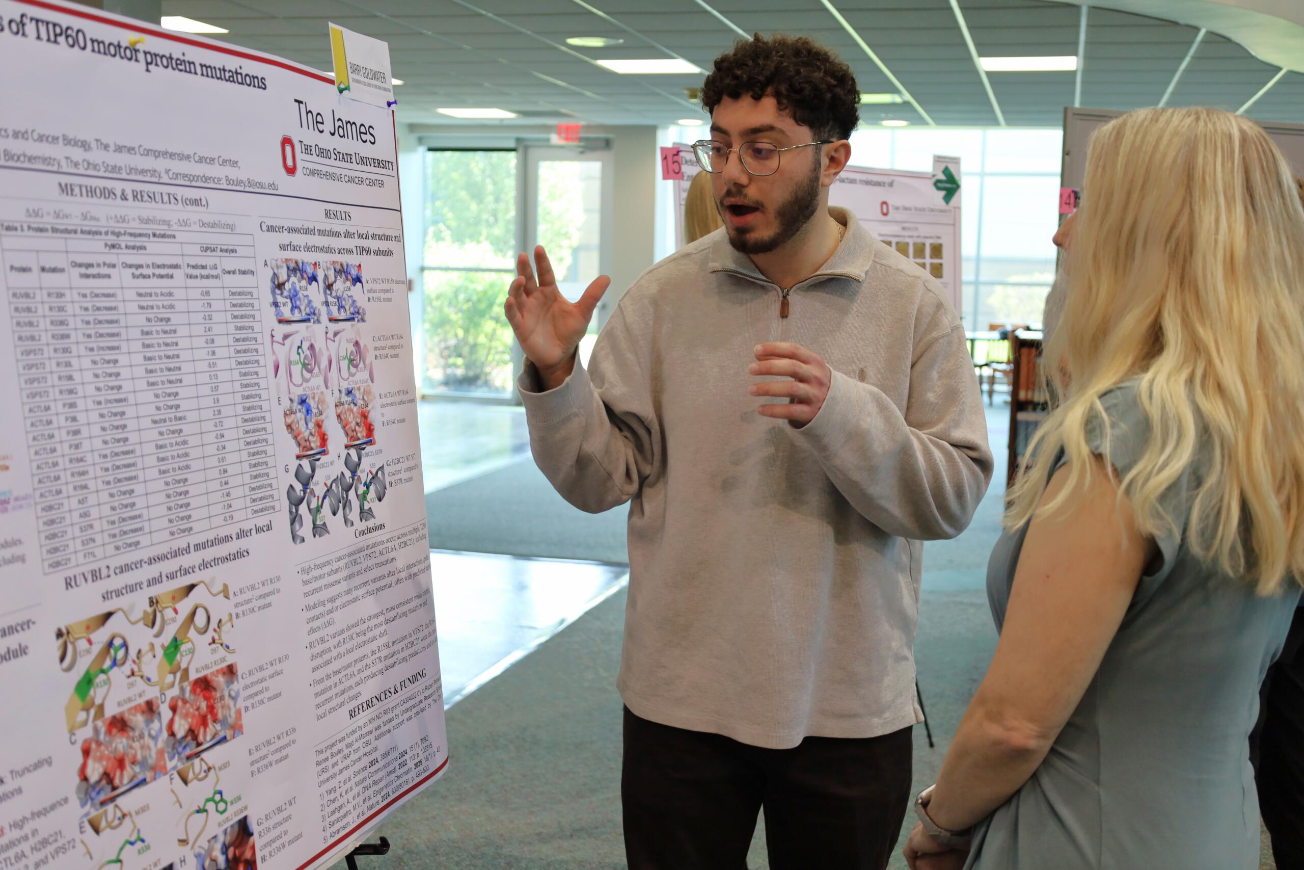 Man explains a scientific poster about cancer-related mutations to a woman at a conference or poster session.