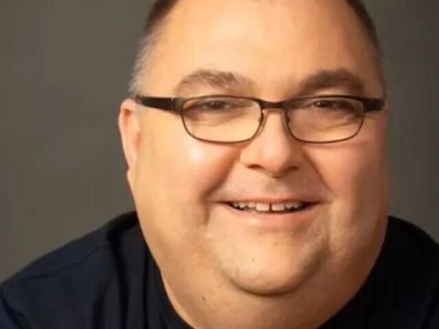Close-up portrait of a smiling man with short hair, glasses, and a dark shirt against a gray background, looking at the camera.