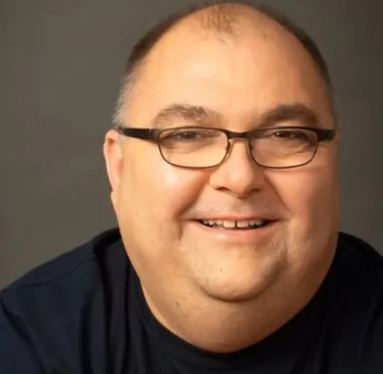 Close-up portrait of a smiling man with short hair, glasses, and a dark shirt against a gray background, looking at the camera.