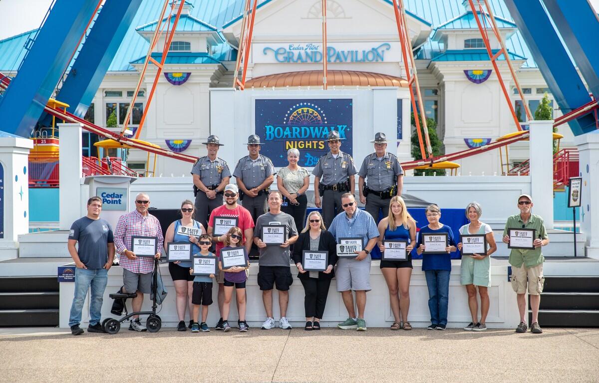 Group of people on a stage at Cedar Point's Grand Pavilion during Boardwalk Nights, holding certificates; officers in uniform stand behind them with a blue-and-white pavilion backdrop.