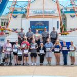 Group of people on a stage at Cedar Point's Grand Pavilion during Boardwalk Nights, holding certificates; officers in uniform stand behind them with a blue-and-white pavilion backdrop.