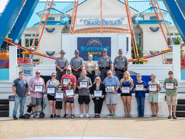 Group of people on a stage at Cedar Point's Grand Pavilion during Boardwalk Nights, holding certificates; officers in uniform stand behind them with a blue-and-white pavilion backdrop.