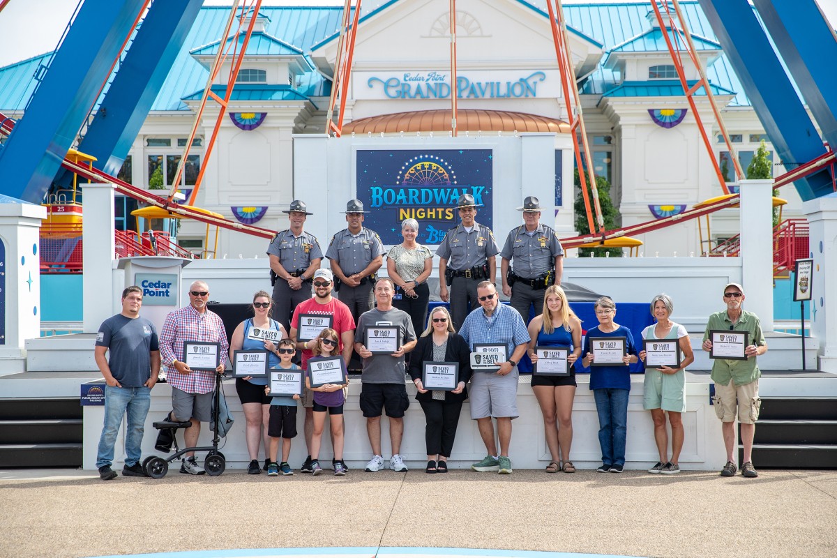 Group of people on a stage at Cedar Point's Grand Pavilion during Boardwalk Nights, holding certificates; officers in uniform stand behind them with a blue-and-white pavilion backdrop.