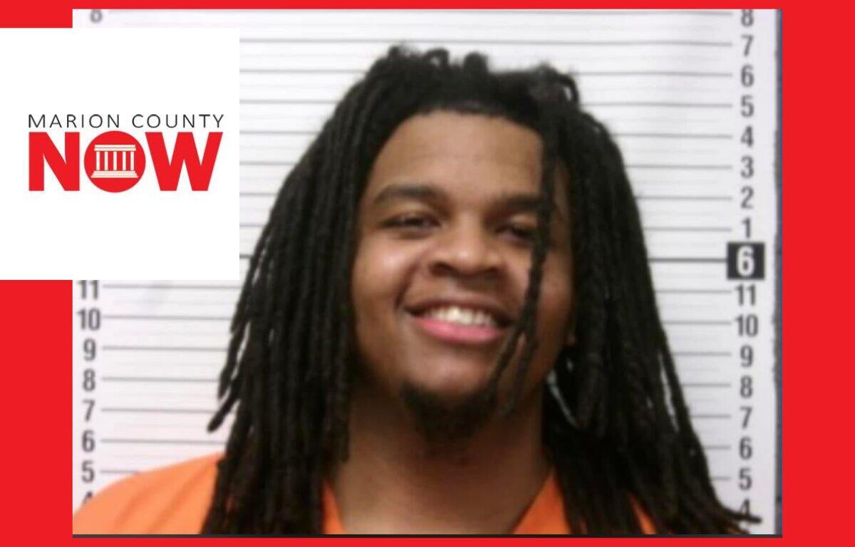 Mugshot of a smiling man with dreadlocks in an orange jumpsuit, height chart behind him.