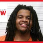 Mugshot of a smiling man with dreadlocks in an orange jumpsuit, height chart behind him.