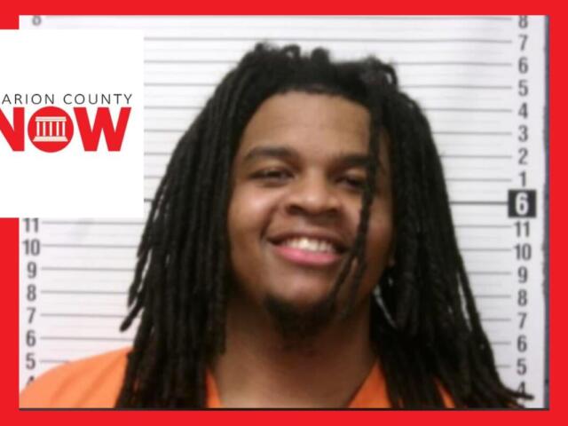 Mugshot of a smiling man with dreadlocks in an orange jumpsuit, height chart behind him.