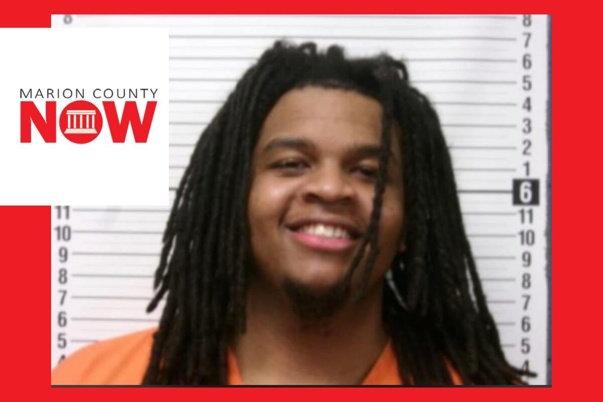 Mugshot of a smiling man with dreadlocks in an orange jumpsuit, height chart behind him.