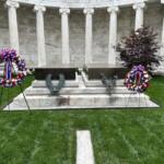 Memorial altar with two wreaths on stands in front of a marble colonnade, on a green lawn.