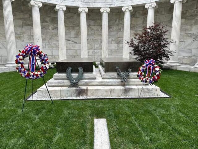 Memorial altar with two wreaths on stands in front of a marble colonnade, on a green lawn.