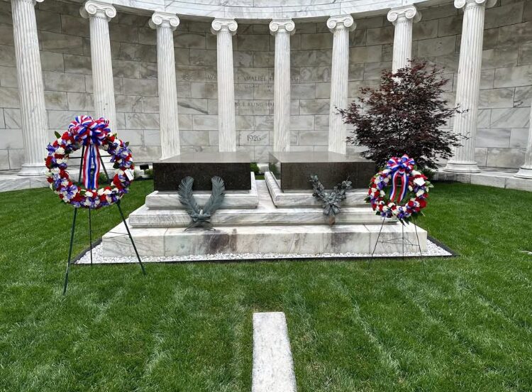 Memorial altar with two wreaths on stands in front of a marble colonnade, on a green lawn.